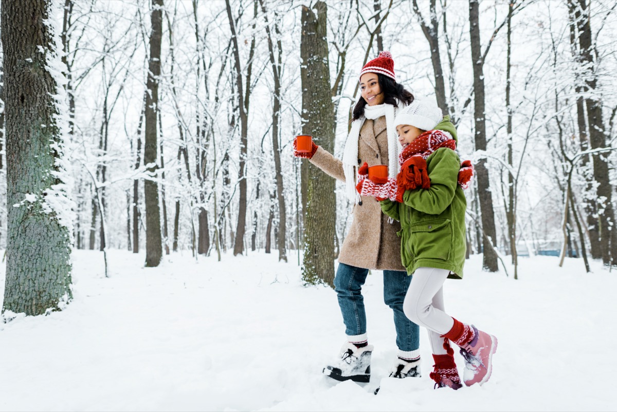 mãe e filha andando e segurando copos vermelhos entre as árvores e olhando para a neve no bosque nevado