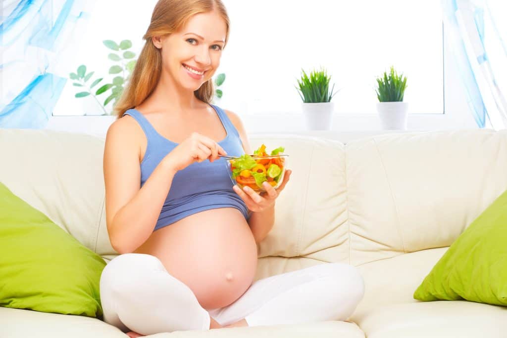 mulher grávida feliz no segundo trimestre come salada de vegetais de comida saudável no sofá em casa