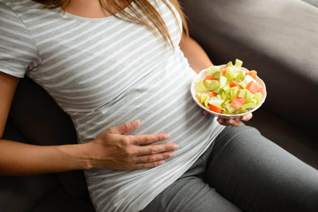 mulher grávida no primeiro trimestre segurando uma tigela de salada verde saudável com alface, abacate e tomate