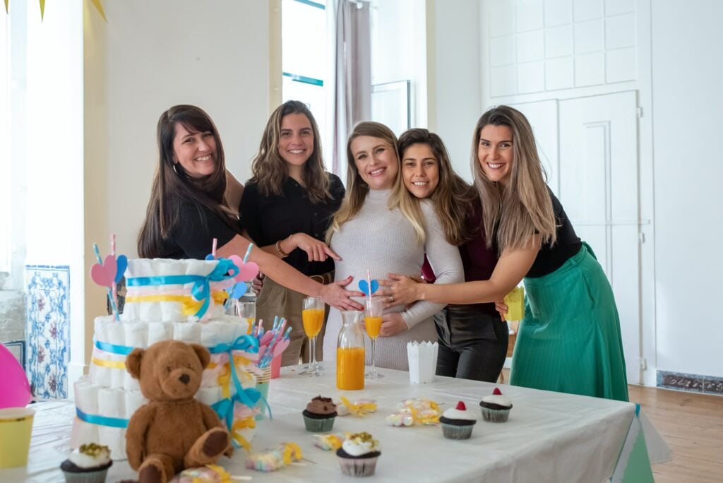 Um grupo de mulheres posa em frente a uma mesa com um bolo de fraldas e um ursinho de pelúcia